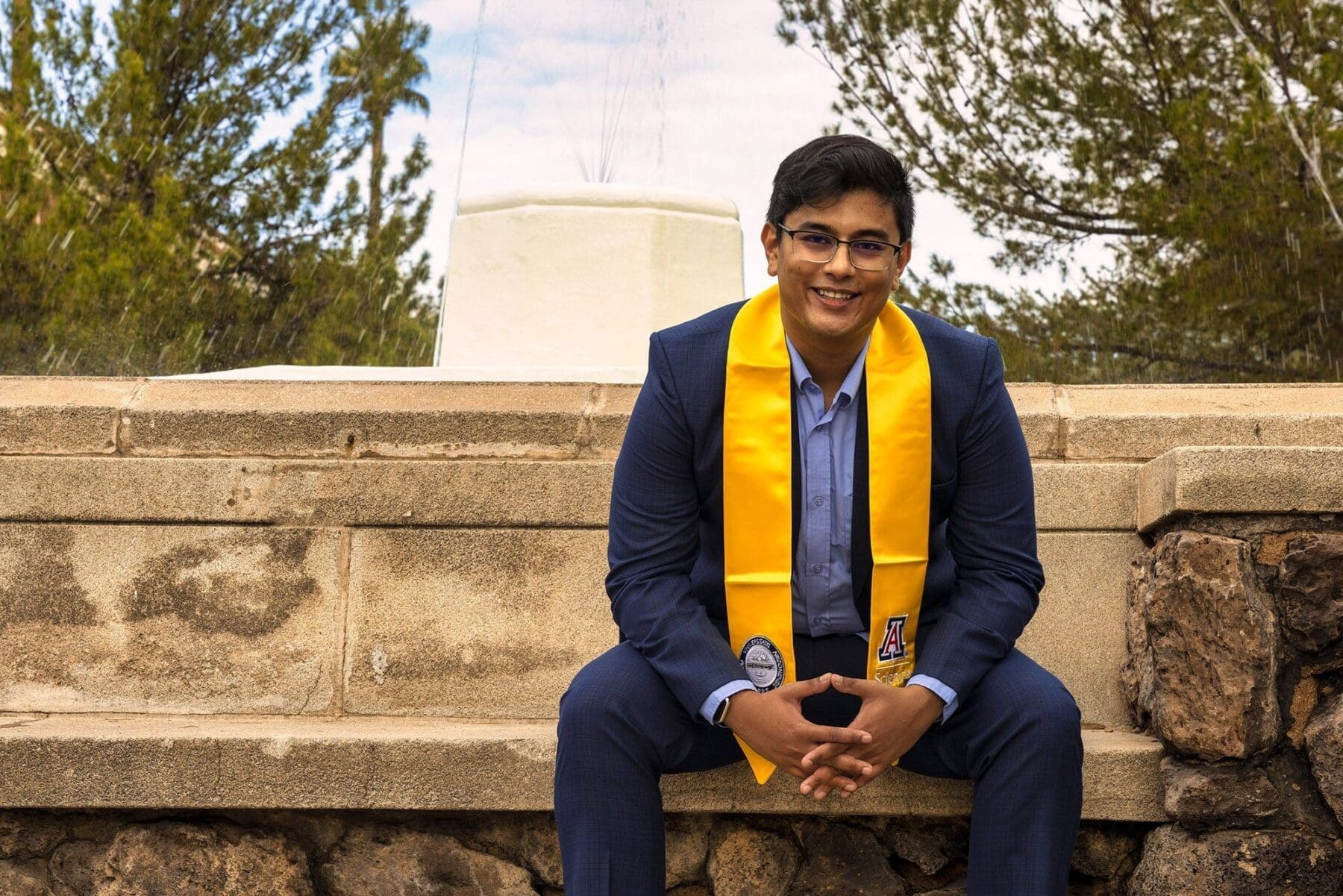 Rehit leans forward and smiles while seated on a low stone wall.