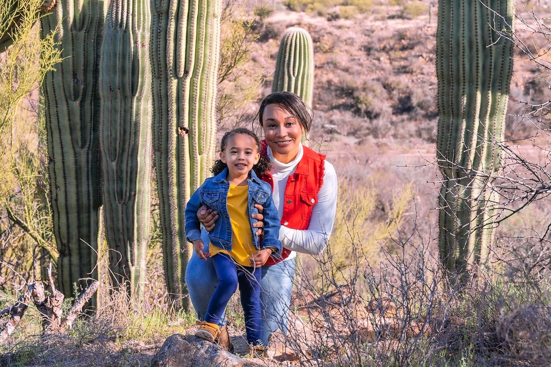 An adult holds a smiling young child among tall saguaros.
