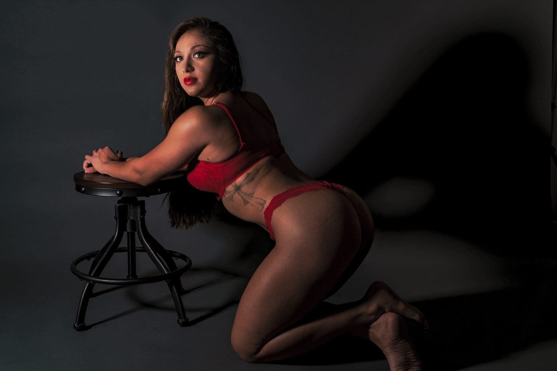 Danni kneels beside a metal stool and looks back toward the camera as her silhouette rises across the wall.