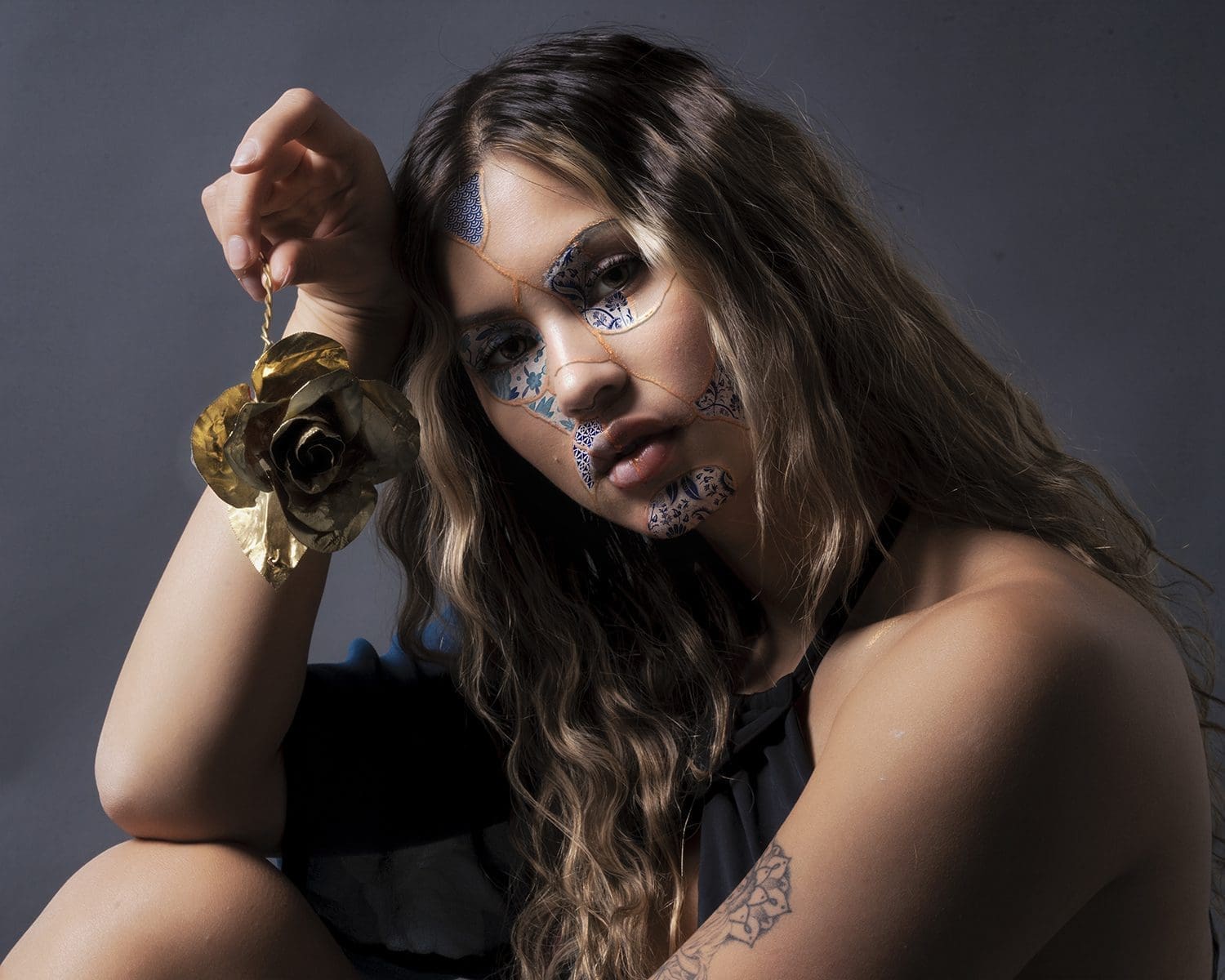 A conceptual studio portrait from the Doll Face series shows the subject resting her head near a metallic gold rose while looking toward the camera.
