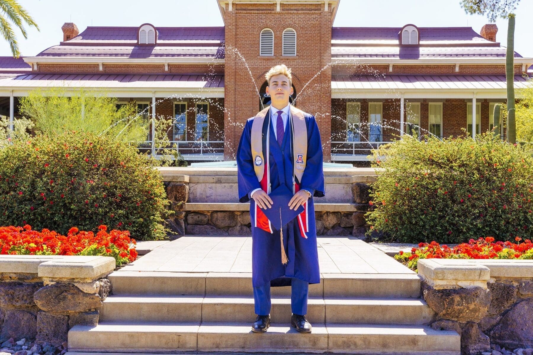 Monty wearing his graduation cap on stone garden steps surrounded by red flowers and shrubs at the University of Arizona