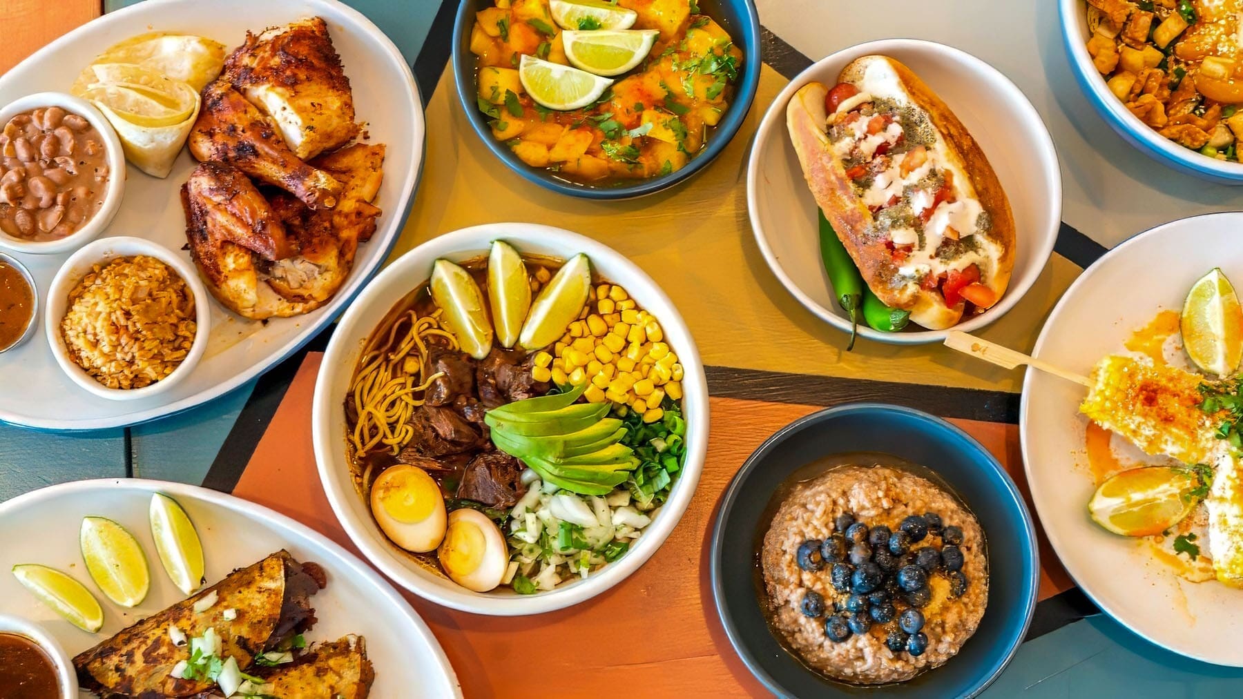 An overhead restaurant photograph presents multiple colorful dishes across a patterned table, including chicken, tacos, bowls, and dessert.