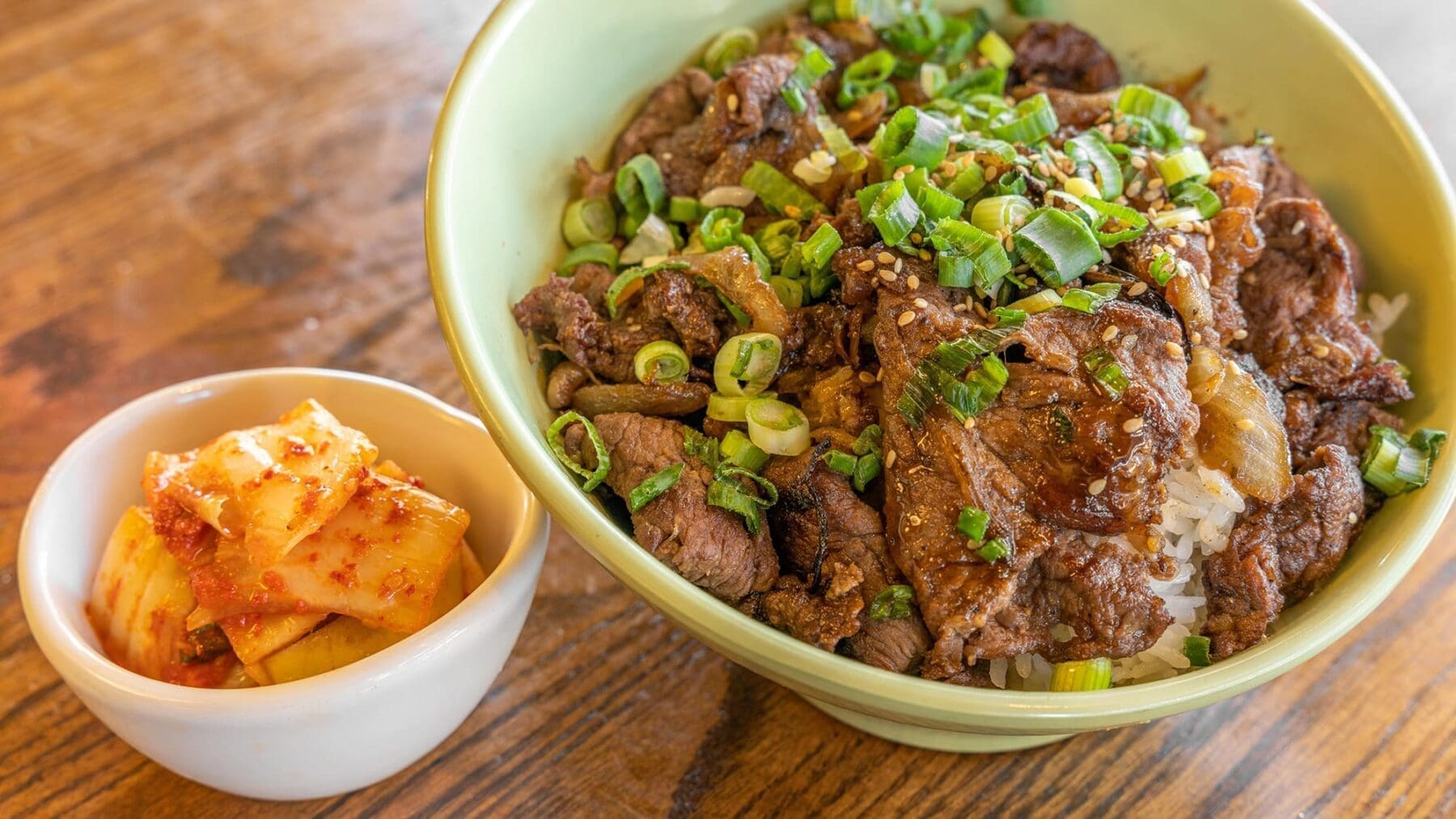 A green bowl holds rice topped with sliced beef, green onion, and sauce, with kimchi served in a small side bowl.